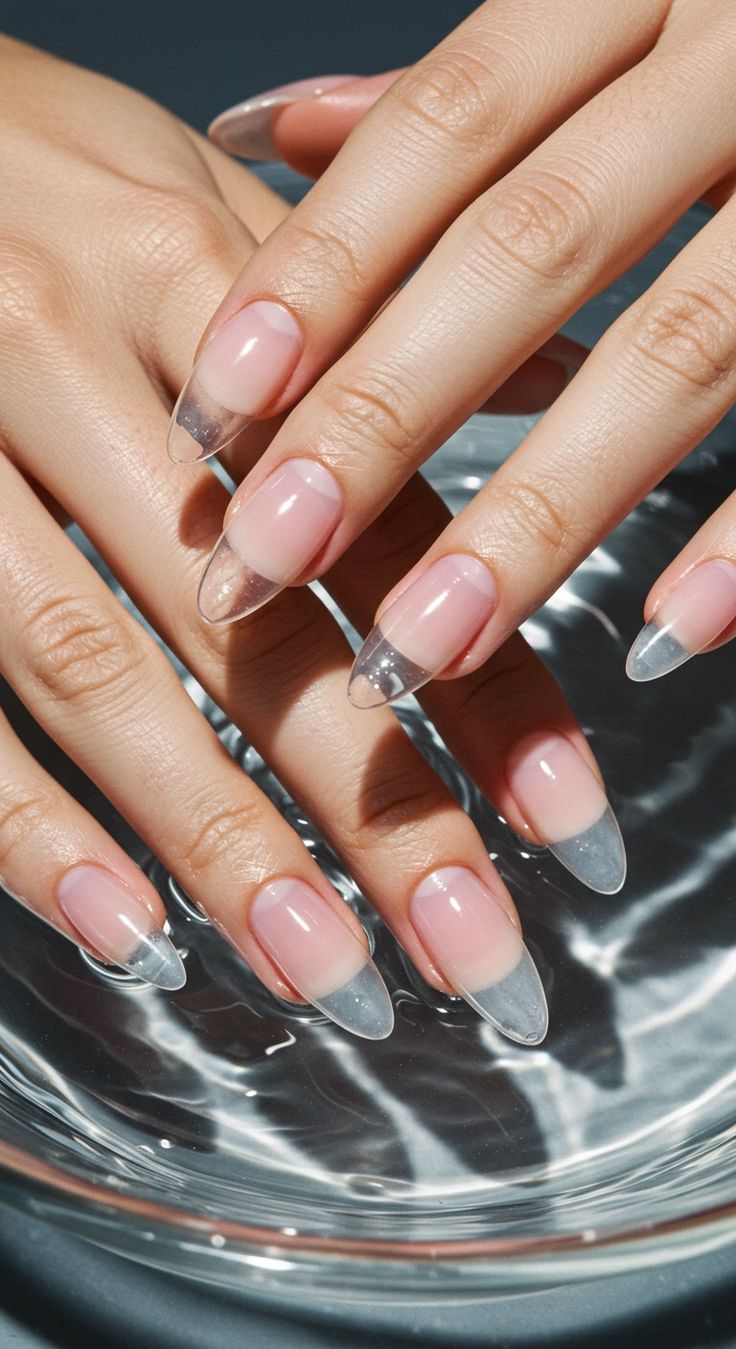 Sheer, natural-look Gel X almond extensions with clear tips, hands resting over water.
